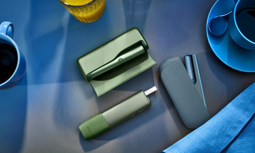 Podgrzewacz IQOS ILUMA leżący na stole obok innych modeli podgrzewaczy IQOS w kolorze Moss Green
