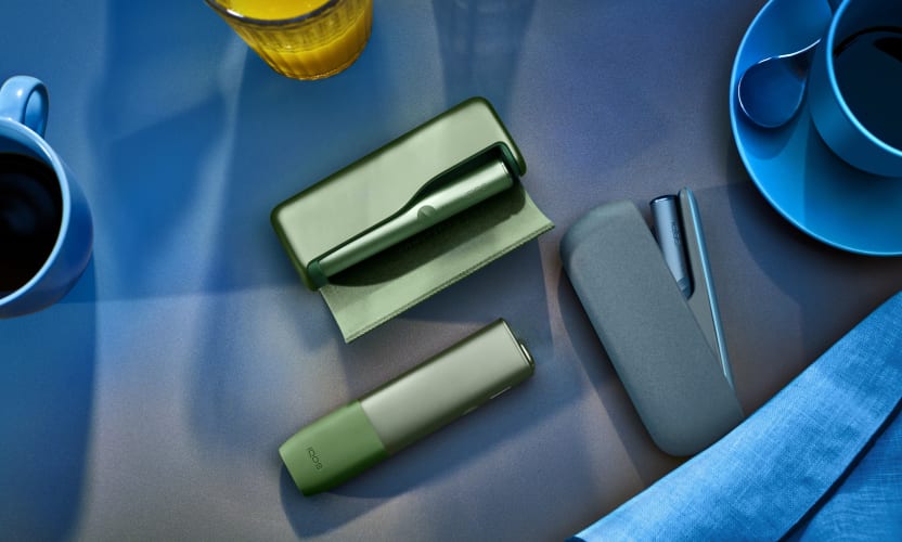 Podgrzewacz IQOS ILUMA leżący na stole obok innych modeli podgrzewaczy IQOS w kolorze Moss Green	