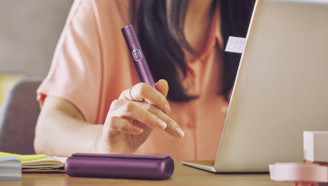 Podgrzewacz IQOS ILUMA i PRIME w dłoni osoby siedzącej przy biurki przy laptopie