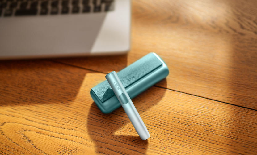 Podgrzewacz IQOS ILUMA i PRIME w kolorze Breeze Blue leżący na stole obok laptopa