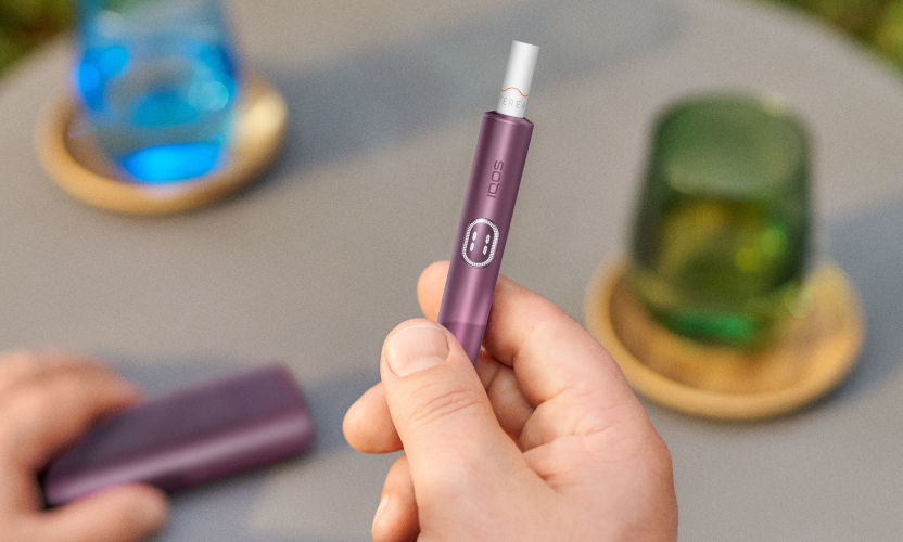 Zbliżenie na trzymanego w dłoni IQOS-a ILUMA i PRIME w kolorze Garnet Red z włożonym wkładem tytoniowym