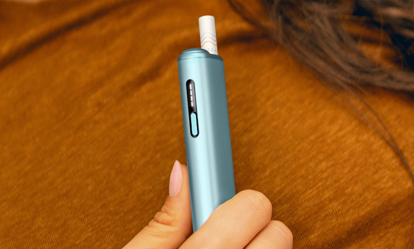 Podgrzewacz tytoniu IQOS ILUMA i ONE w kolorze Breeze Blue, z wsuniętym wkładem tytoniowym, trzymany w dłoni