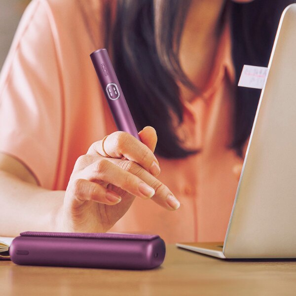 Kobieta siedząca przed laptopem, trzymająca w ręce podgrzewacz IQOS ILUMA i PRIME w kolorze Garnet Red