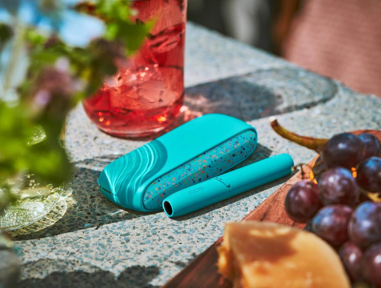 The WE Edition IQOS 3 DUO limited edition charger in turquoise with flecks of color on the door cover next to the holder beside a drink and plate of fruit.​